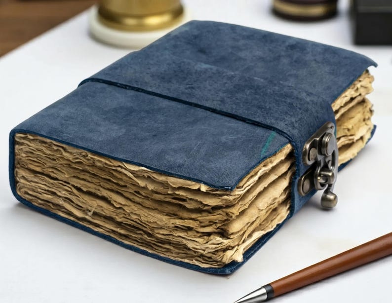 May include: A rectangular journal with a deep blue textured cover and a metal latch closure. The journal contains thick, deckled-edge pages with a light tan, aged appearance. A brown pen is positioned in the lower right corner of the frame.