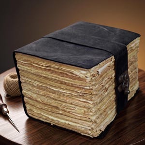 May include: A thick, rectangular book with a matte black leather cover and a metal latch closure. The pages are numerous, uneven, and have a light tan, aged appearance. A small ball of twine and a metal awl with a wooden handle sit nearby.