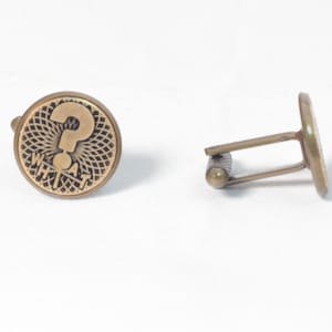 Puede incluir: Un par de gemelos dorados. Cada gemelo presenta un diseño circular con un signo de interrogación en el centro, rodeado por un patrón radial y la palabra "WHAT". Los gemelos tienen un cierre de barra en T.