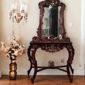 May include: A dark wood console table with ornate carvings and a large, ornately framed mirror. The table has a gold finish and sits on a light brown wood floor.