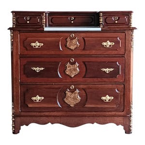 Puede incluir: Una cómoda de madera oscura con tres cajones y una sección superior con pequeños cajones y un espacio central abierto. La cómoda presenta herrajes dorados y detalles decorativos en forma de escudo en los frentes de los cajones. La madera tiene un rico tono marrón rojizo.