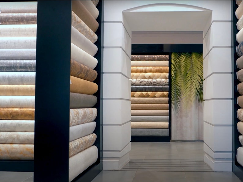 K&ouml;nnte beinhalten: Eine Wand mit Regalen, die mit Rollen von Tapeten in verschiedenen Farben und Mustern gef&uuml;llt sind. Die Regale sind in einem Raster angeordnet und die Tapeten sind ordentlich gestapelt. Die Tapeten sind in verschiedenen Texturen und Farben, darunter Beige, Braun, Grau und Wei&szlig;.