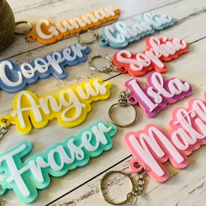 May include: A selection of personalised name keyrings in various colours. Each keyring displays a name in a white, cursive font, set against a coloured background. Keyring colours include orange, blue, yellow, pink, and green, with silver key rings attached.