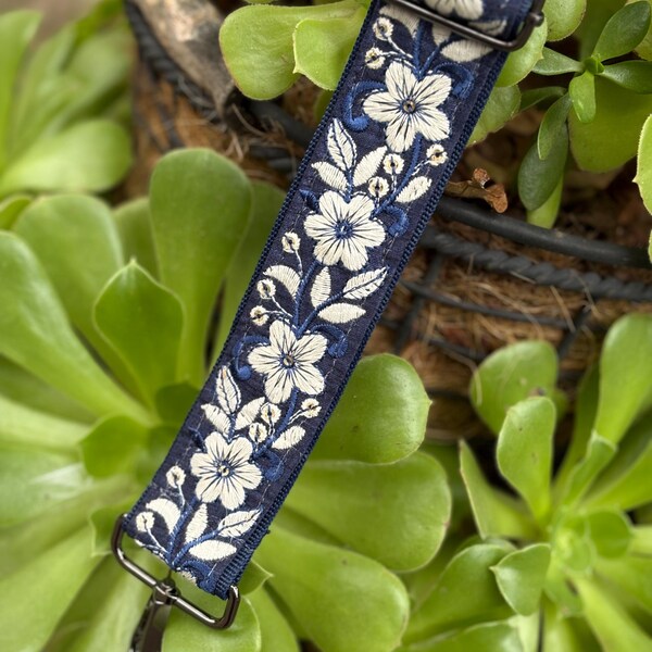 Op de afbeelding: Een marineblauwe band met witte bloemenborduurwerk. De band heeft een herhalend patroon van witte bloemen en bladeren, met een metalen sluiting en hardware. De band staat tegen een achtergrond van groene vetplanten.
