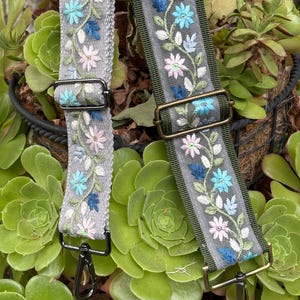 May include: Two embroidered bag straps with floral patterns. One strap is gray with black hardware, the other is olive green with brass hardware. Both straps feature pink, blue, and white flower embroidery.