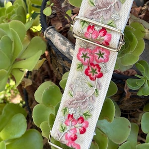 May include: A cream-colored fabric bag strap featuring an intricate floral embroidery pattern in shades of pink, red, and green with metallic silver accents. The strap includes polished silver-tone metal hardware, including a sliding adjuster and a swivel clip.
