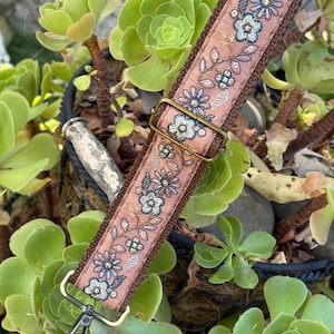 May include: A brown fabric bag strap featuring an embroidered floral pattern in shades of blue, white, and gold. The strap includes an adjustable antique brass metal slider and a swivel lobster clasp hook at the end for secure attachment.