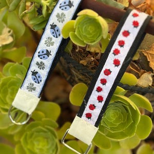 Può includere: Due cinturini da polso in tessuto con fettuccia nera e nastri bianchi sovrapposti. Un cinturino presenta un motivo di coccinelle azzurre e trifogli grigi, mentre l'altro mostra una fila di piccole coccinelle rosse. Entrambi hanno anelli a D in metallo argentato.