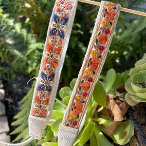 Pode incluir: Duas correias decorativas tecidas com bordados florais em laranja, azul e amarelo. As correias são de cor creme com um tecido texturizado e são exibidas em um fundo de folhagem verde. As correias são provavelmente para uma câmera ou bolsa.