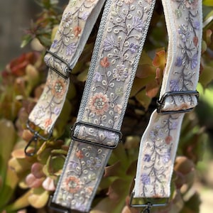 May include: Cream-colored embroidered bag straps with floral patterns in shades of peach, purple, and green. The straps have black metal hardware and are designed for attaching to a bag. The floral embroidery adds a decorative touch.