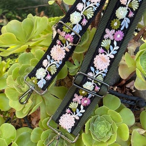 May include: Two adjustable bag straps featuring a black fabric base with intricate floral embroidery in shades of pink, light blue, and green. Each strap includes silver-toned metal hardware, including rectangular sliders and swivel lobster clasps.