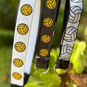 Pode incluir: Três chaveiros com tema de voleibol. Um é branco com bolas de voleibol amarelas, outro é preto com bolas de voleibol amarelas e o terceiro é branco com bolas de voleibol pretas. Cada chaveiro tem um anel de metal.