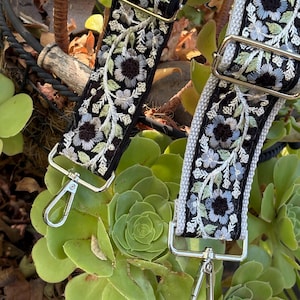 May include: A black bag strap featuring intricate floral embroidery in shades of light blue, white, and sage green. The strap includes adjustable silver-toned metal sliders and two silver-toned swivel lobster claw clasps at the ends.