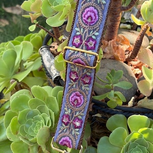 May include: A replacement bag strap featuring a light blue fabric base with intricate floral embroidery in shades of magenta, purple, and green. The strap includes gold-toned metal hardware, including an adjustable slider and a swivel lobster clasp.