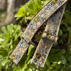 Puede incluir: Dos correas bordadas para bolsos con motivos florales en tonos azules, verdes y beige. Las correas tienen herrajes metálicos y están colocadas sobre un fondo de follaje verde. Las correas están anguladas entre sí.