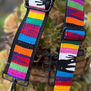 Op de afbeelding: Twee zwarte geweven tasriemen met een levendig, kleurrijk gestreept patroon. De riemen hebben een regenboog aan kleuren, waaronder rood, oranje, geel, groen, blauw en paars. Elke riem heeft zwarte metalen hardware en een draaibare sluiting.