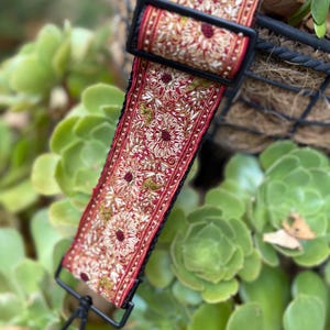 Pode incluir: Uma alça decorativa com um padrão floral em vermelho, dourado e verde. A alça tem uma fivela preta e um fecho de metal preto. A alça é provavelmente para uma bolsa ou carteira.