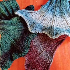 May include: Three crocheted scarves in different color gradients. The scarves are made with a textured stitch and have a scalloped edge. The colors are green, blue, and burgundy.