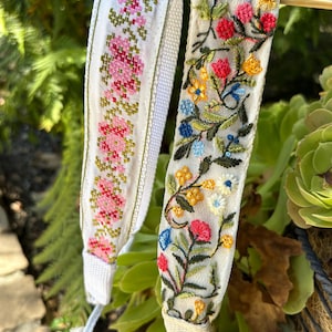 Puede incluir: Dos correas de tela blanca con bordados florales. Una correa presenta flores rosas de punto de cruz, mientras que la otra muestra un diseño floral bordado colorido con elementos rojos, amarillos, azules y verdes. Las correas cuelgan sobre un fondo verde.