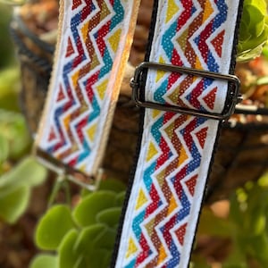 May include: Two patterned bag straps with a zigzag design in red, yellow, blue, and brown on a white background. The straps have metal fixings and are set against a backdrop of green foliage.