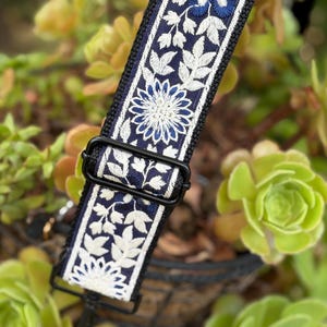 May include: A replacement bag strap featuring a dark navy blue fabric base with intricate white and blue floral embroidery. The strap includes a black metal adjustable slider buckle and a black metal swivel clasp at the end for secure attachment.