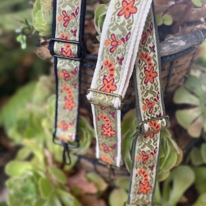 May include: Close-up of three decorative bag straps with floral embroidery. The straps feature orange flowers and green leaves on a cream, black, and olive green background. Each strap has metal hardware for attachment.