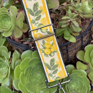 May include: A decorative bag strap featuring a lemon and leaf pattern on a cream background with yellow borders. The strap has silver-toned metal hardware, including a rectangular buckle and a swivel clasp. The strap is set against a backdrop of green succulents.