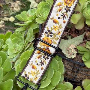 May include: Cream-colored bag strap with a floral pattern in shades of yellow, brown, and green. The strap has a dark brown border and black metal hardware, including a clasp and a rectangular buckle. The strap is set against a background of green succulent plants.