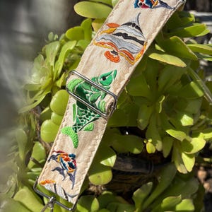 Może przedstawiać: Beżowy pasek do aparatu z haftowanymi wzorami wodnymi w różnych kolorach. Pasek posiada zielonego żółwia, kolorowe ryby oraz srebrną metalową klamrę i zapięcie. Pasek jest prezentowany na tle zielonych roślin sukulentów.