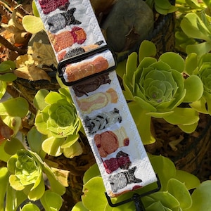 May include: A white fabric bag strap featuring a repeating embroidered pattern of small dogs in various colors including orange, brown, gray, and black. The strap includes black metal hardware, an adjustable sliding buckle, and a swivel lobster clasp.