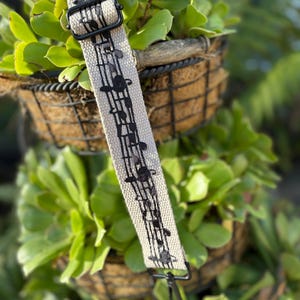 以下が含まれることがあります： 黒い楽譜が印刷されたベージュのギターストラップ。ストラップには黒い金属製のバックルと黒い金属製のクリップが付いています。