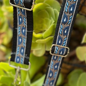 May include: Close-up of two black straps with a decorative pattern in blue, light blue, and tan. The straps have silver-colored metal hardware, including adjustable buckles and swivel clasps. The background is a blurred view of green foliage.