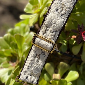 May include: Cream-colored embroidered strap with a dark brown backing and gold-colored metal hardware. The strap features a floral design with intricate detailing. The background is a blurred view of green foliage.