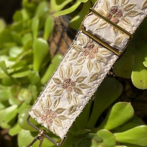 Pode incluir: Close-up de uma alça de bolsa creme com bordados florais em tons de ouro e rosa. A alça apresenta ferragens metálicas douradas, incluindo fivelas de ajuste retangulares e um fecho giratório. O fundo é uma vista desfocada de plantas suculentas verdes.