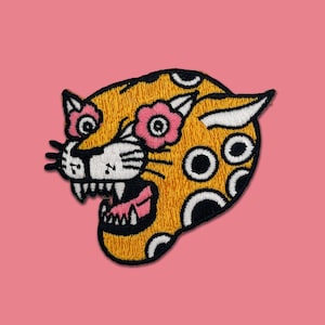 May include: Embroidered patch featuring a stylized jaguar head with a floral design. The jaguar is yellow with black spots and outlines, white teeth, and pink flowers. The patch is set against a pink background.