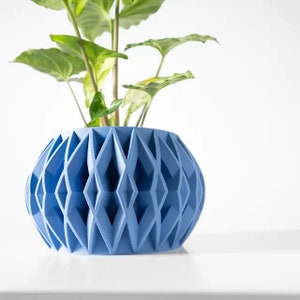 Könnte beinhalten: Hellblauer Blumentopf mit geometrischem Muster, der eine grüne Pflanze mit Blättern enthält. Der Topf hat ein rautenförmiges Design und besteht aus einem festen Material. Die Pflanze hat große, herzförmige Blätter. Der Topf steht auf einer weißen Oberfläche.
