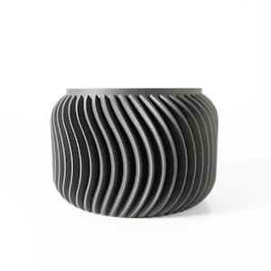 Könnte beinhalten: Ein grauer, zylindrischer Behälter mit einem einzigartigen, wellenartigen Design. Die Außenseite weist vertikale, gebogene Linien auf, die eine strukturierte Oberfläche erzeugen. Der Behälter besteht wahrscheinlich aus Metall oder einem ähnlichen Material.