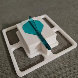 May include: A white plastic dart holder with a teal dart and a white square base. The dart has a teal flight and a clear shaft. The holder has a square frame with rectangular cutouts.