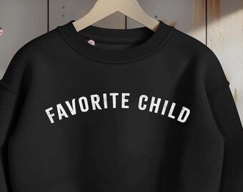 May include: Black sweatshirt with the text "FAVORITE CHILD" in white, hanging on a wooden hanger. The sweatshirt is displayed against a light wood background with a shelf and decorative floral accents.