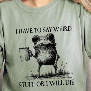May include: Sage green t-shirt with a graphic of a frog holding a mug, with the text "I HAVE TO SAY WEIRD STUFF OR I WILL DIE". The shirt is worn with blue jeans.