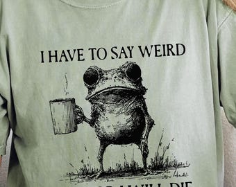May include: Sage green t-shirt with a graphic of a frog holding a mug, with the text "I HAVE TO SAY WEIRD STUFF OR I WILL DIE". The shirt is worn with blue jeans.