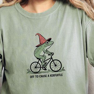 May include: Sage green t-shirt featuring a whimsical illustration of a frog riding a bicycle and wearing a wizard hat. The text "OFF TO CAUSE A KERFUFFLE" is printed below the image. The shirt is made of a soft material.