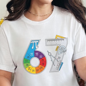 May include: White t-shirt with a colorful graphic design featuring the numbers "67" and a pi symbol. The design includes a rainbow-colored circle with numbers and mathematical tools.