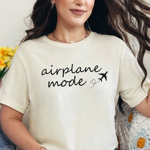 K&ouml;nnte beinhalten: Eine Frau tr&auml;gt ein cremefarbenes T-Shirt mit dem Text "airplane mode" und einer kleinen Flugzeuggrafik. Sie sitzt auf einem Sofa mit einem blauen Kissen und einer geh&auml;kelten Decke.