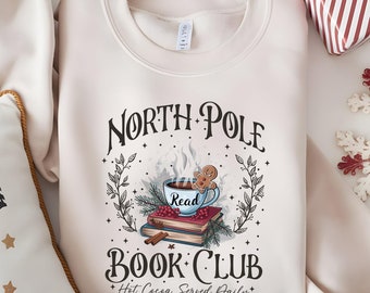 May include: A beige sweatshirt with a graphic design of a mug of hot cocoa with a gingerbread man, books, and a wreath. The text on the sweatshirt reads "North Pole Book Club *Hot Cocoa Served Daily*"