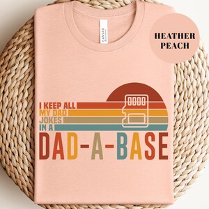 May include: Heather peach-colored t-shirt with the text "I KEEP ALL MY DAD JOKES IN A DAD-A-BASE" in a retro design. The design includes a sunset graphic with a data storage symbol.