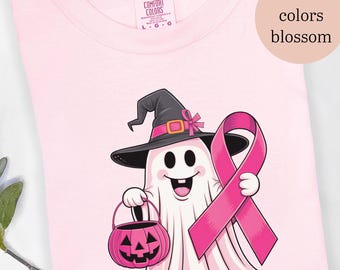 May include: Pink t-shirt featuring a cartoon ghost wearing a witch hat and holding a pink ribbon and a jack-o'-lantern. The shirt has the words "comfort colors blossom" on it.