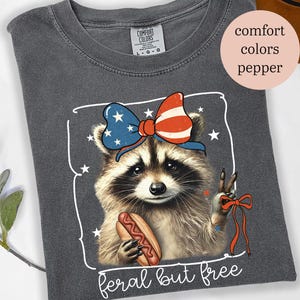 Op de afbeelding: Donkergrijs T-shirt met een wasbeer die een patriottische strik draagt en een hotdog vasthoudt. De tekst "feral but free" staat onder de afbeelding van de wasbeer.