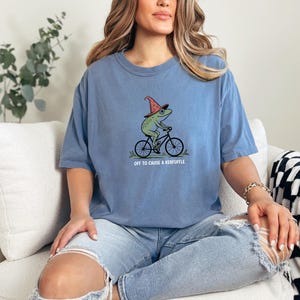 May include: A light blue t-shirt featuring a green frog riding a bicycle and wearing a red wizard hat. The text "OFF TO CAUSE A KERFUFFLE" is printed below the frog. The shirt is made of a soft material.