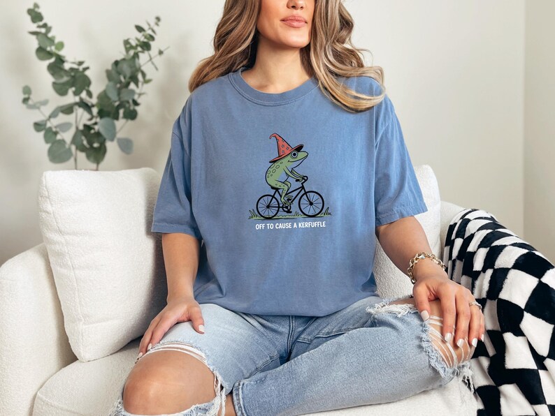 May include: A light blue t-shirt featuring a green frog riding a bicycle and wearing a red wizard hat. The text "OFF TO CAUSE A KERFUFFLE" is printed below the frog. The shirt is made of a soft material.